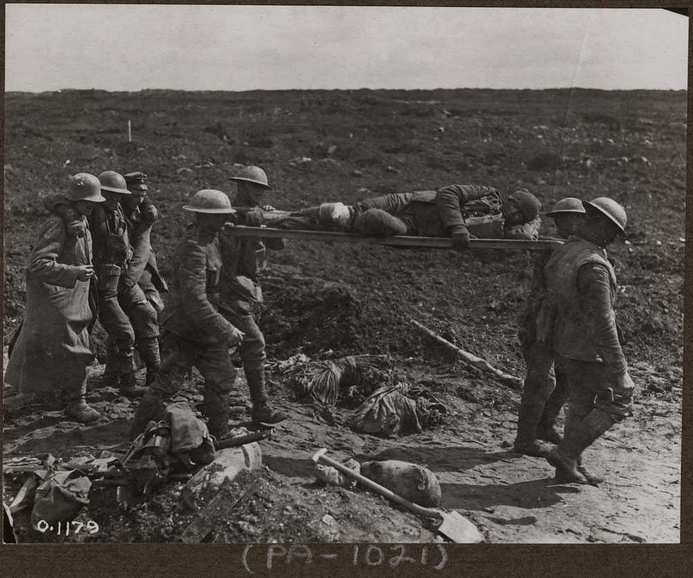 Black and white photograph. A muddy battlefield. Four men in military uniforms and helmets carry a man on a stretcher on their shoulders. Three men follow behind them, the man in the centre supported by the other two.  Debris litters the ground.